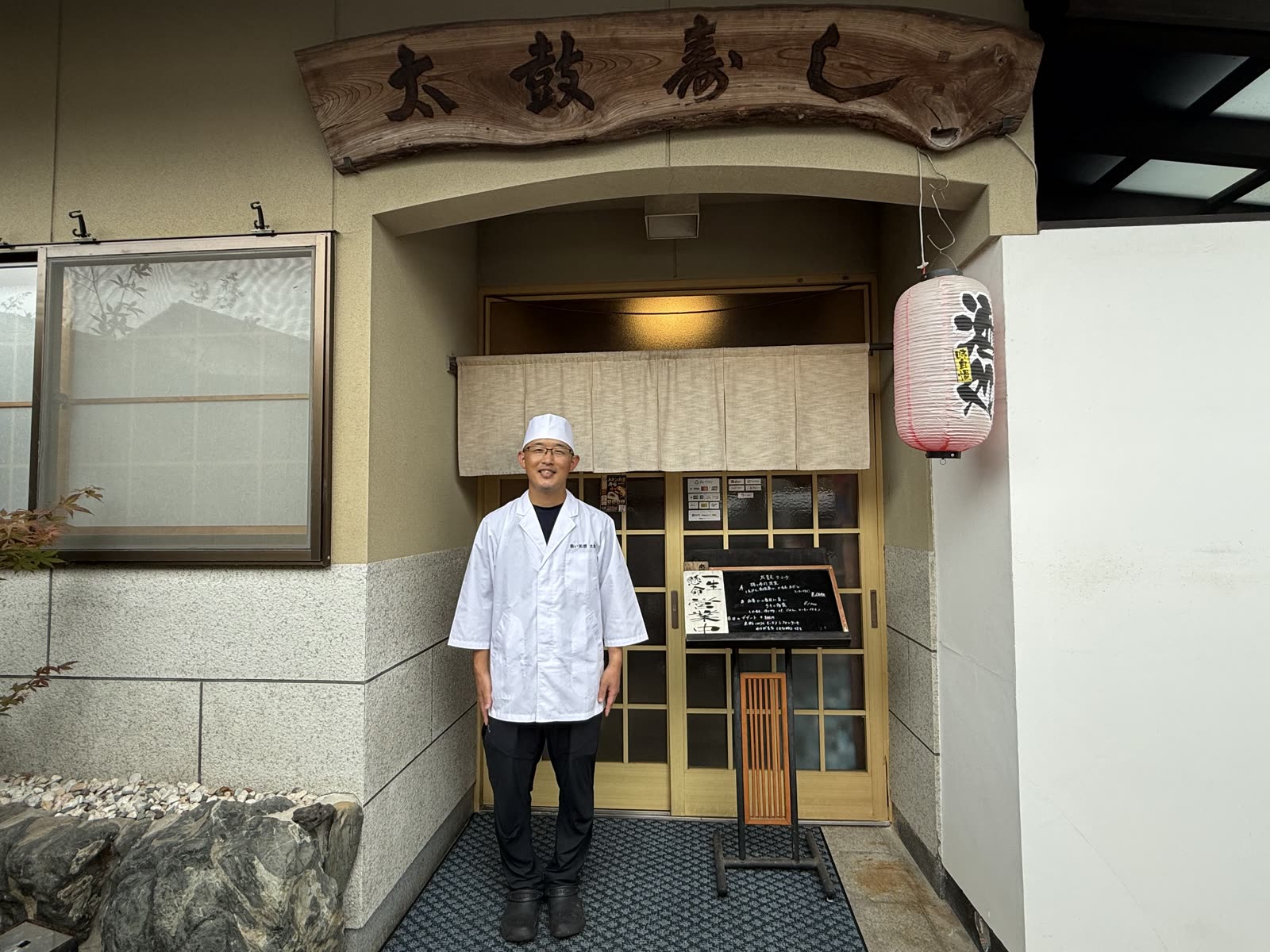 「太鼓寿し」の木彫りの看板と、店主の大石昌平